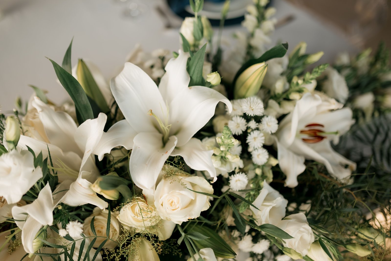 A white floral arrangement with lilies, roses, and small white flowers on a white tablecloth.