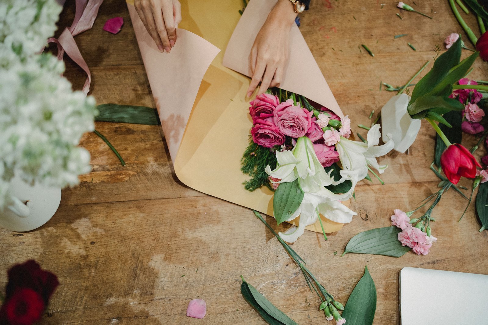 Hands wrap pink and white flowers in peach and yellow paper on a wooden table.