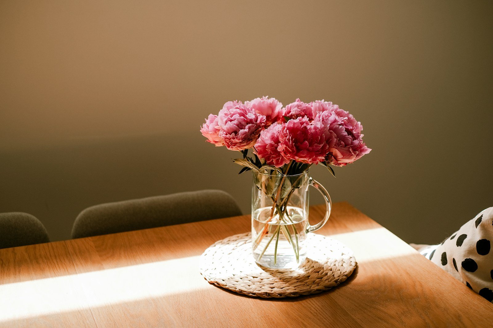 Pink peonies in a glass pitcher sit on a woven mat on a wooden table.