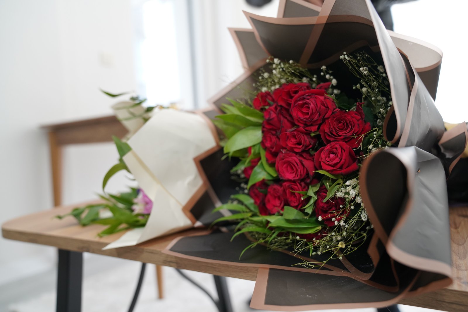 A bouquet of red roses and baby's breath in dark and light wrapping sits on a wooden table.
